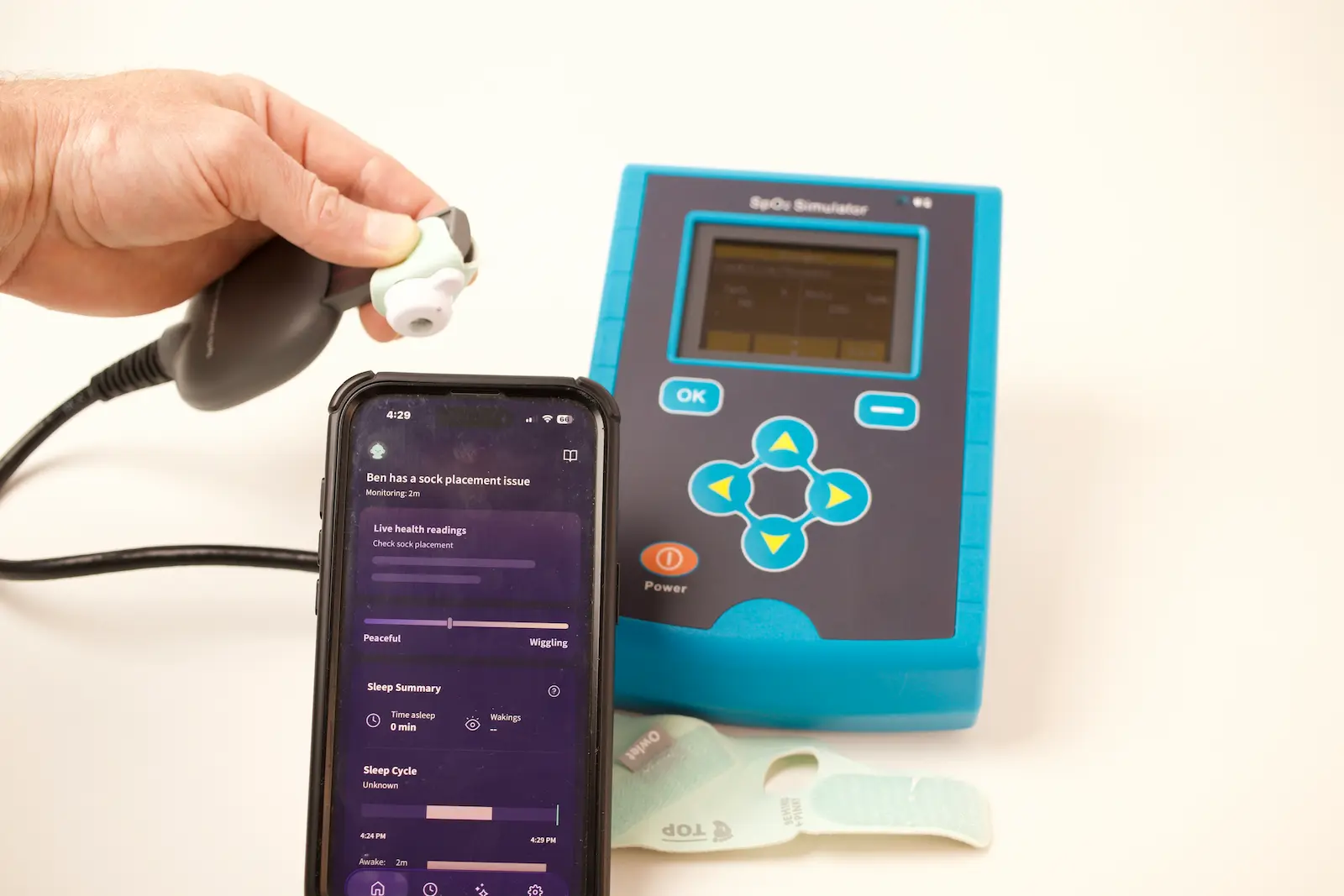 Hand holding pulse oximeter sensor near SpO2 simulator device with Owlet baby monitor app showing sock placement alert on sma