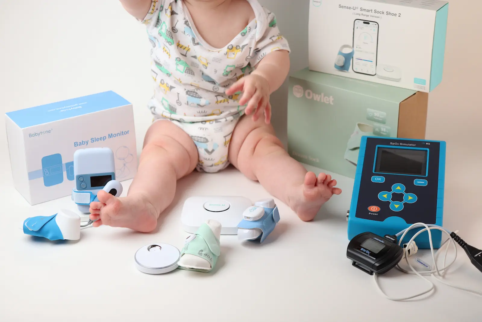 Baby surrounded by infant sleep monitors including Owlet, Sense-U Smart Sock, and Babytone Baby Sleep Monitor devices