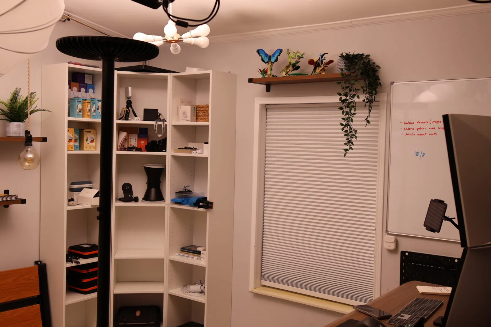 Organized home office with white shelving unit storing tech gadgets, whiteboard with notes, window blinds, and decorative pla