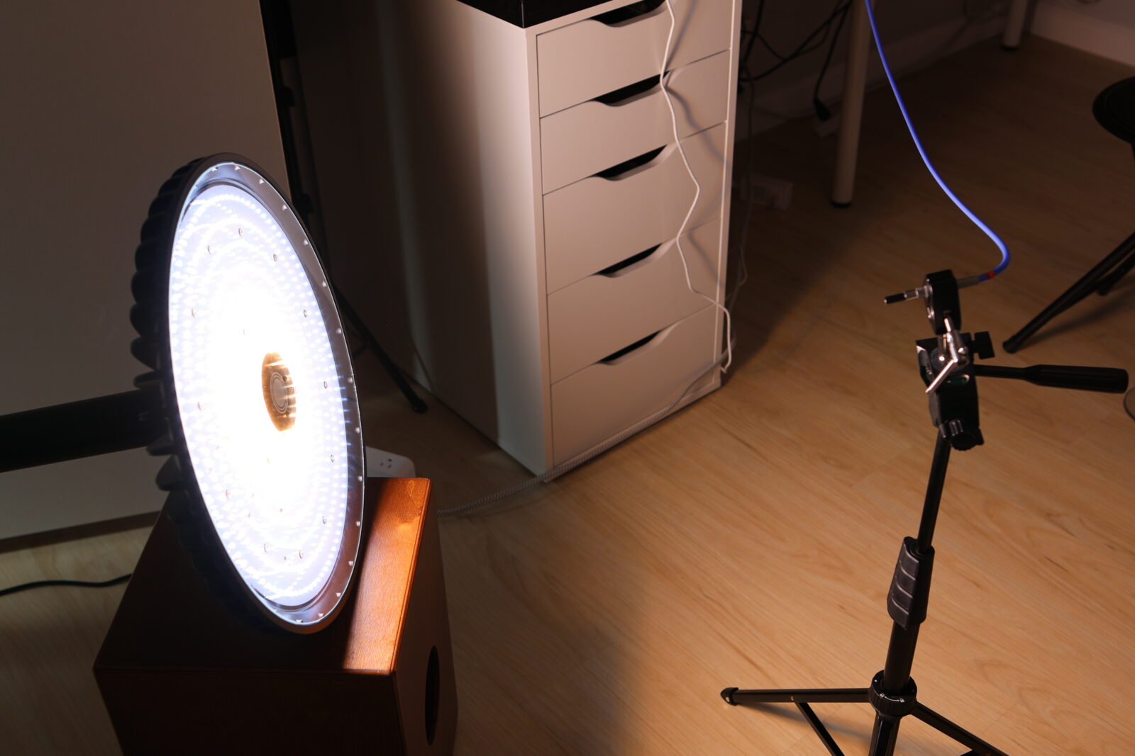 Large circular LED studio light glowing brightly on a wooden speaker, beside a white drawer unit and light stand