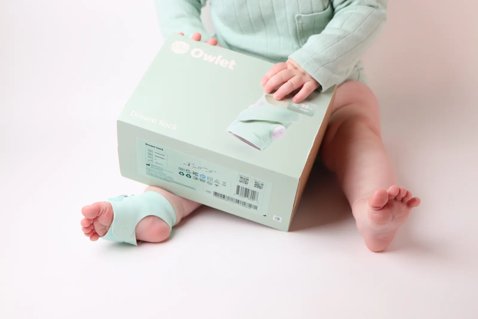 Baby wearing Owlet Dream Sock on foot while holding mint green product box, seated on white background