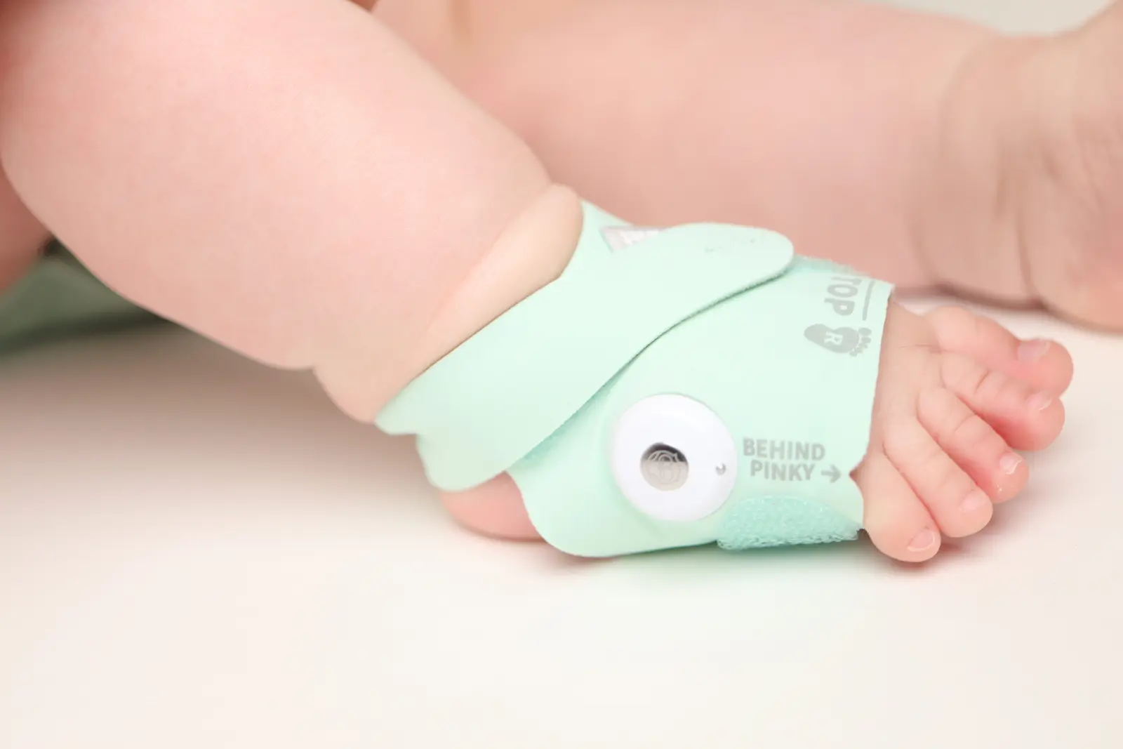 Baby foot wearing a mint green pulse oximeter sensor wrap labeled Behind Pinky for infant oxygen monitoring