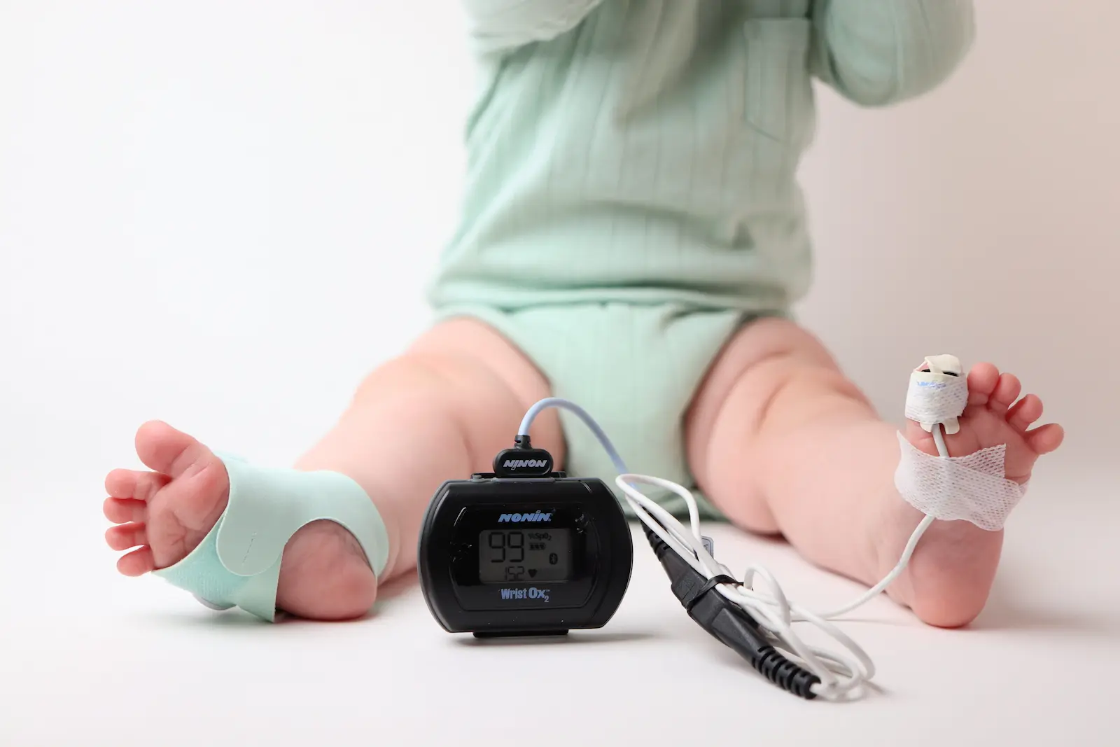 Baby sitting with Nonin WristOx2 pulse oximeter showing 99% SpO2, sensor wrapped on foot with medical gauze