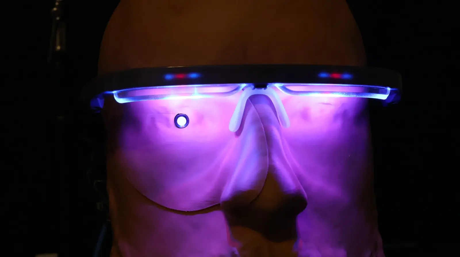 Mannequin head wearing glowing blue LED glasses with purple UV light illuminating a silicone face mask in darkness