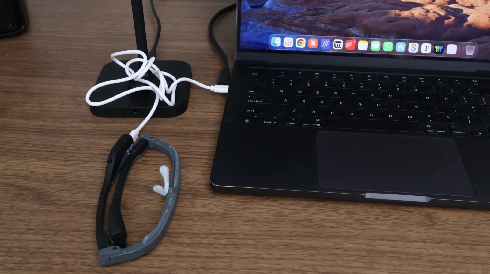 AR smart glasses connected via white USB cable to a MacBook Pro on a wooden desk with apps visible in Dock