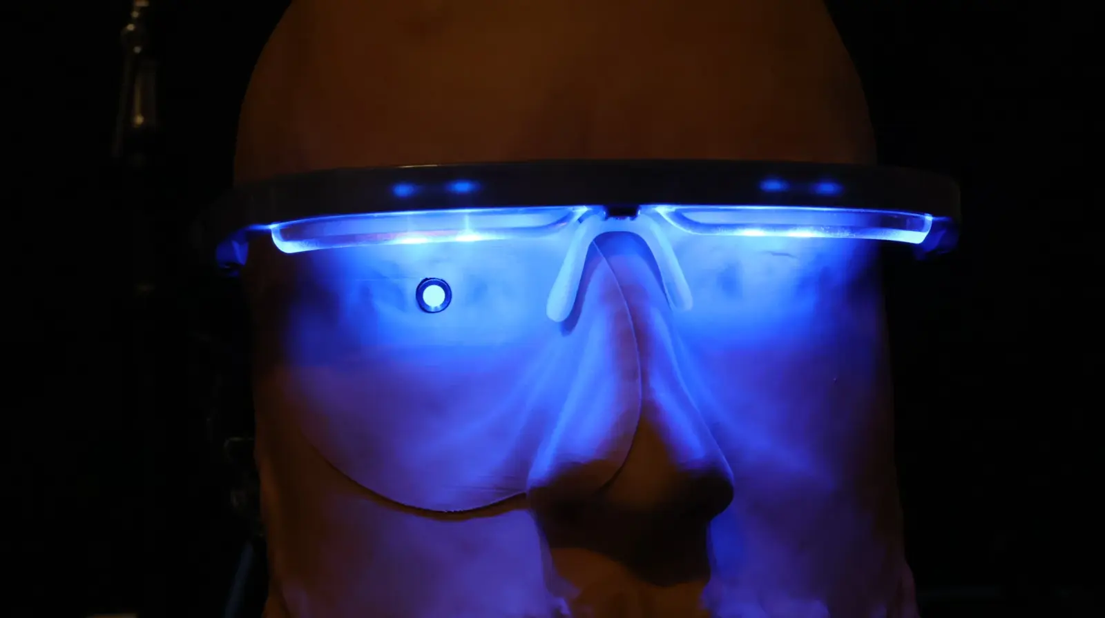 Bald person wearing futuristic blue LED-illuminated smart glasses in a dark dramatic setting