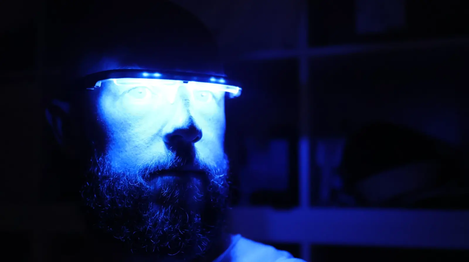Bearded man wearing futuristic blue LED smart glasses in a dark cyberpunk-style setting