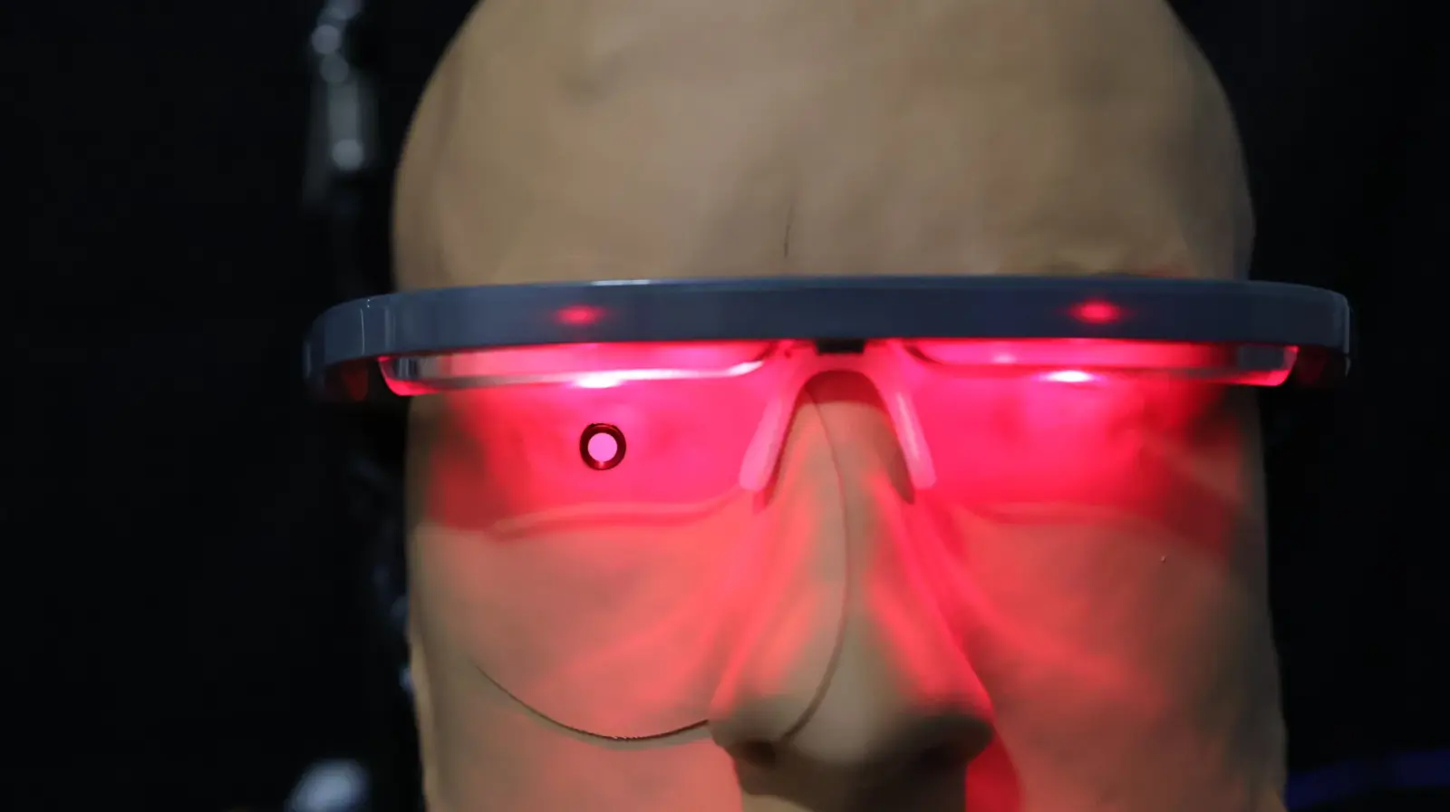 Bald person wearing glowing red LED light therapy glasses against dark background