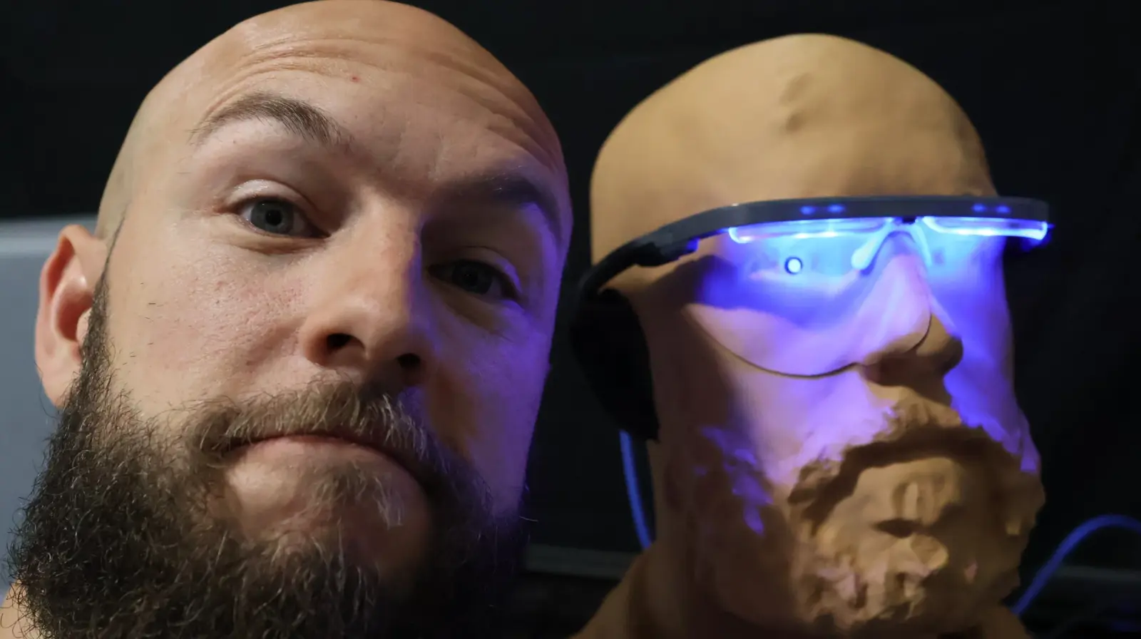 Bald man with beard posing beside a realistic 3D-printed sculpture of his own face wearing glowing blue LED glasses