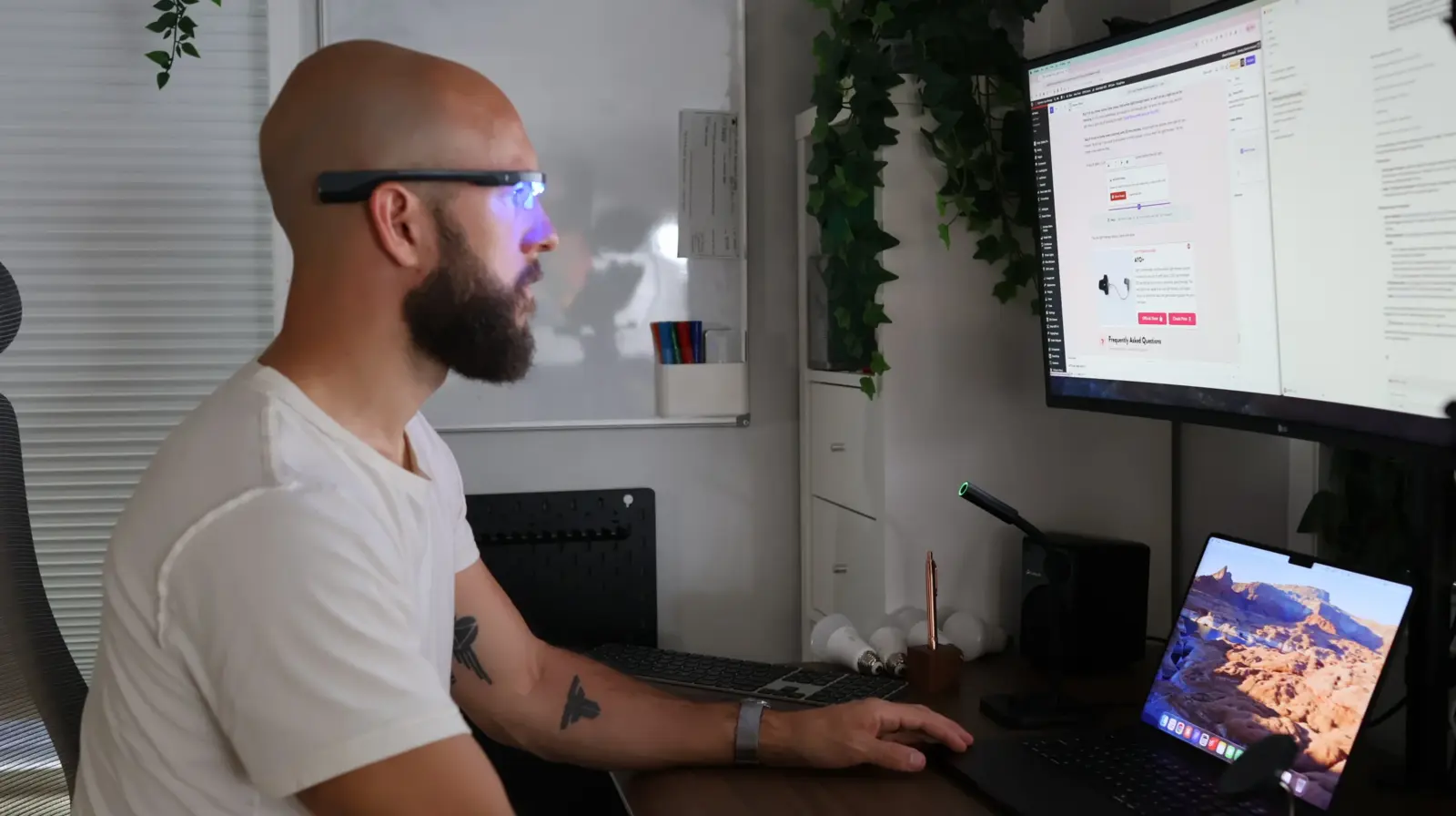 Bald man with beard wearing smart AR glasses working at multi-monitor desk setup with MacBook and LG display