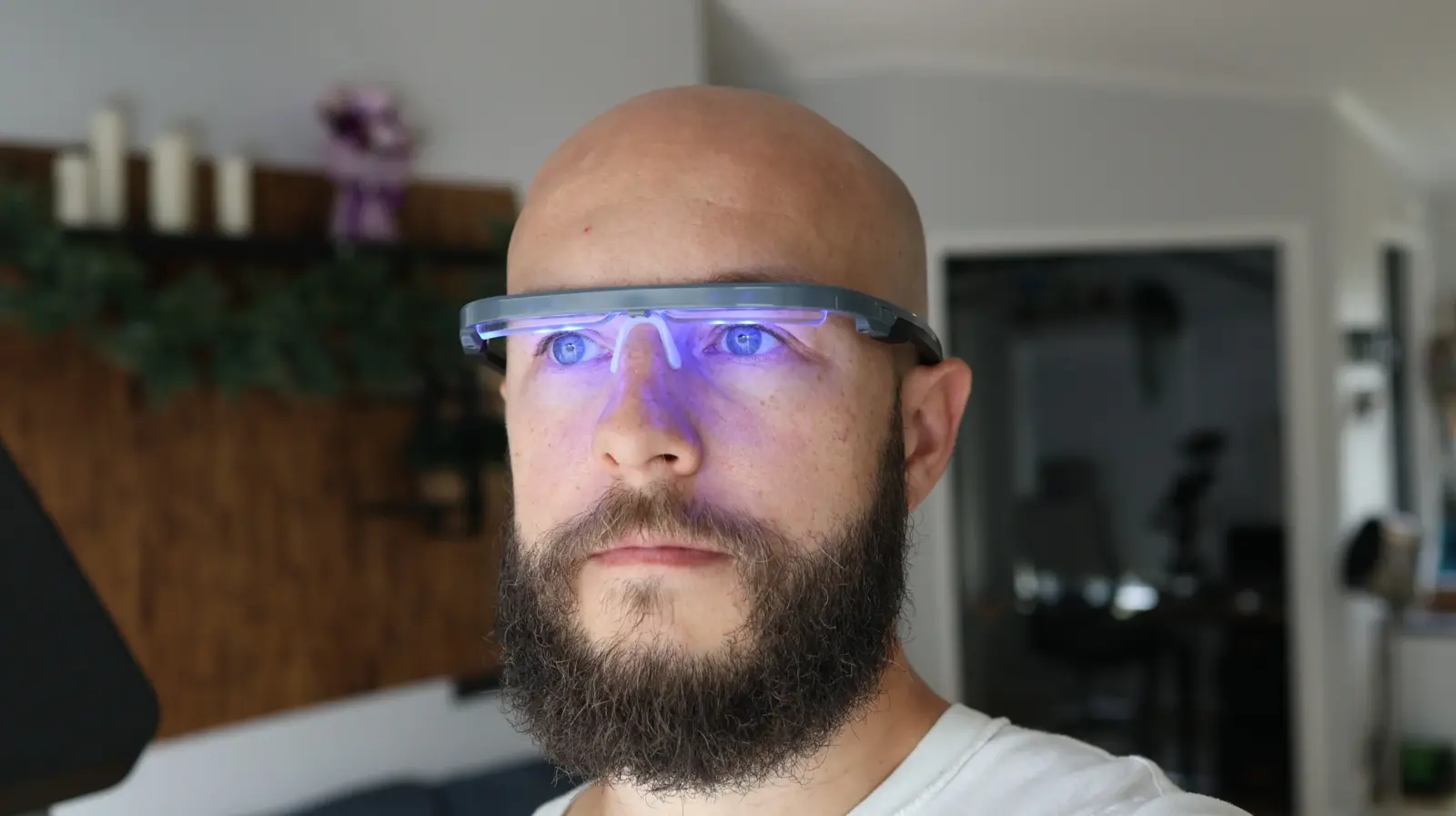 Bald bearded man wearing glowing blue LED light therapy glasses indoors