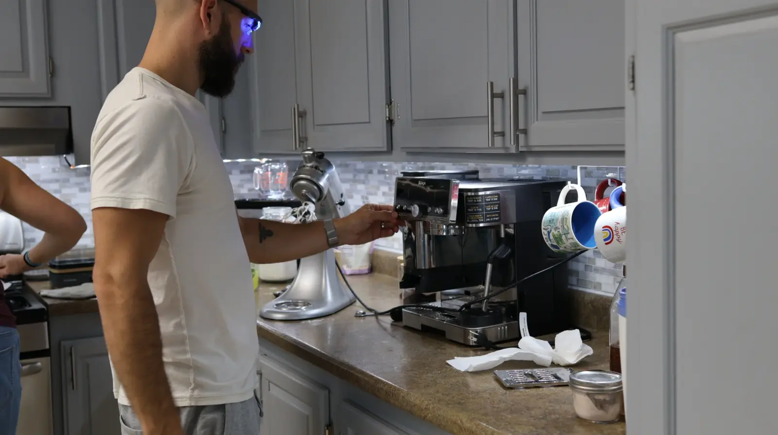 Bearded man in white t-shirt operating a Ninja espresso machine on a kitchen counter with mugs and a KitchenAid mixer nearby