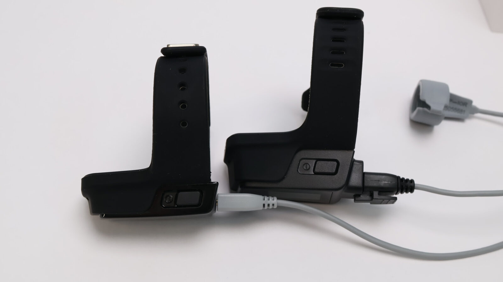 Two black fitness tracker wristbands connected to charging cables with a pulse oximeter sensor on white background