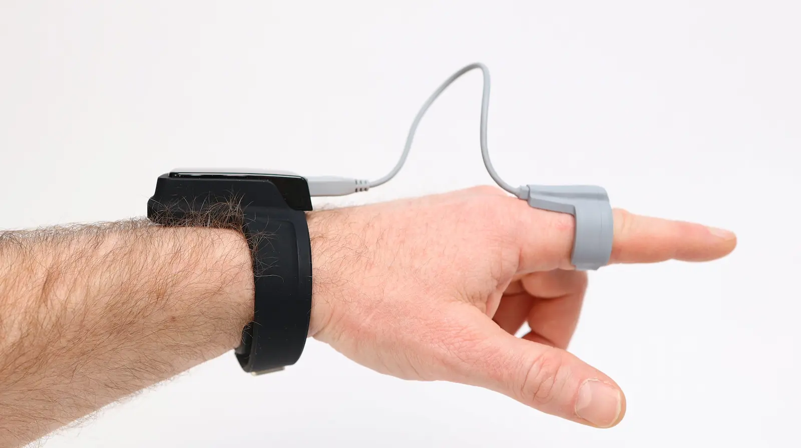 Hand wearing a black wrist-worn health monitor connected by cable to a gray pulse oximeter finger sensor