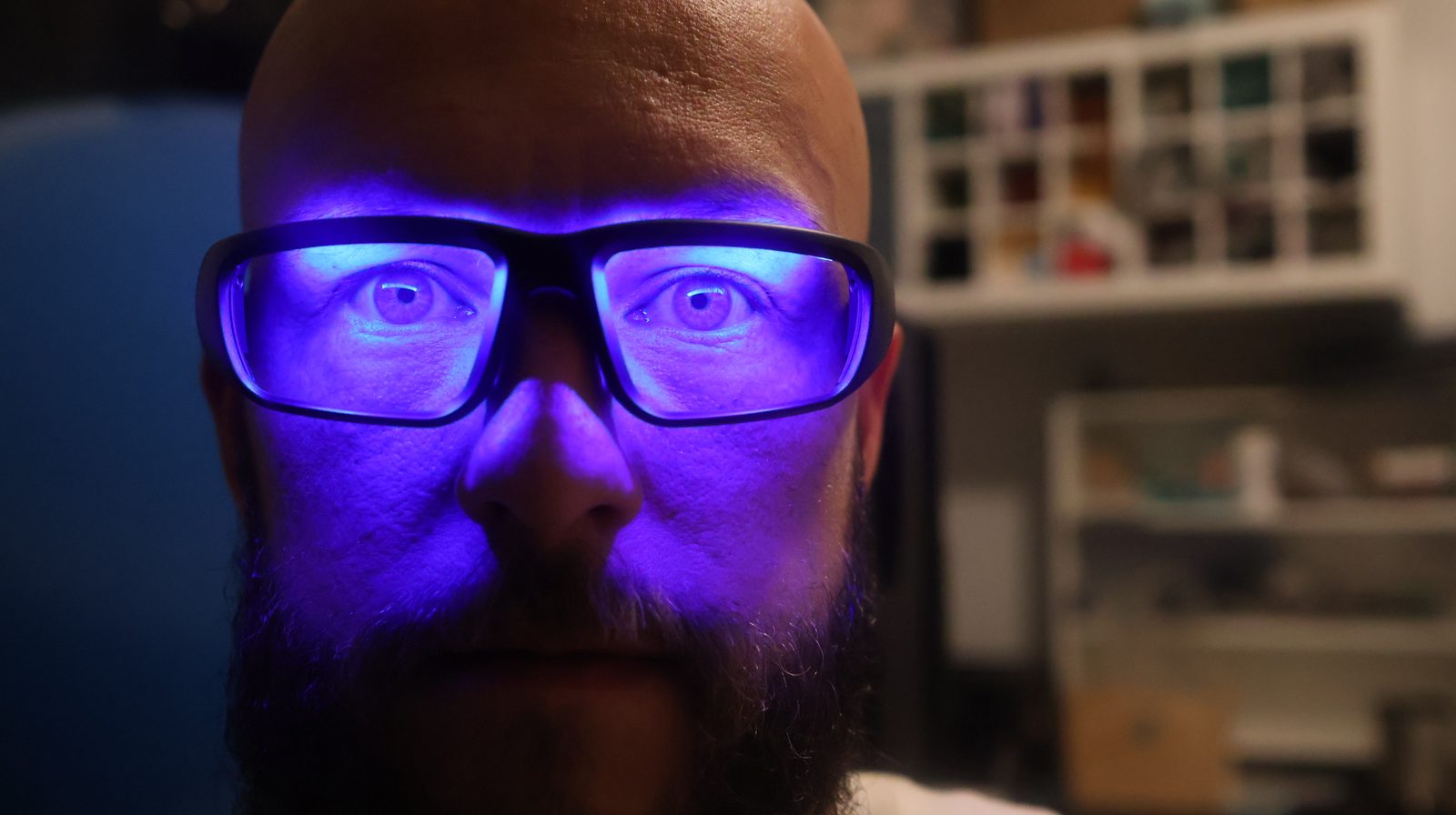 Bald bearded man wearing glowing blue-lit glasses in a dark room, illuminated by vivid purple-blue light