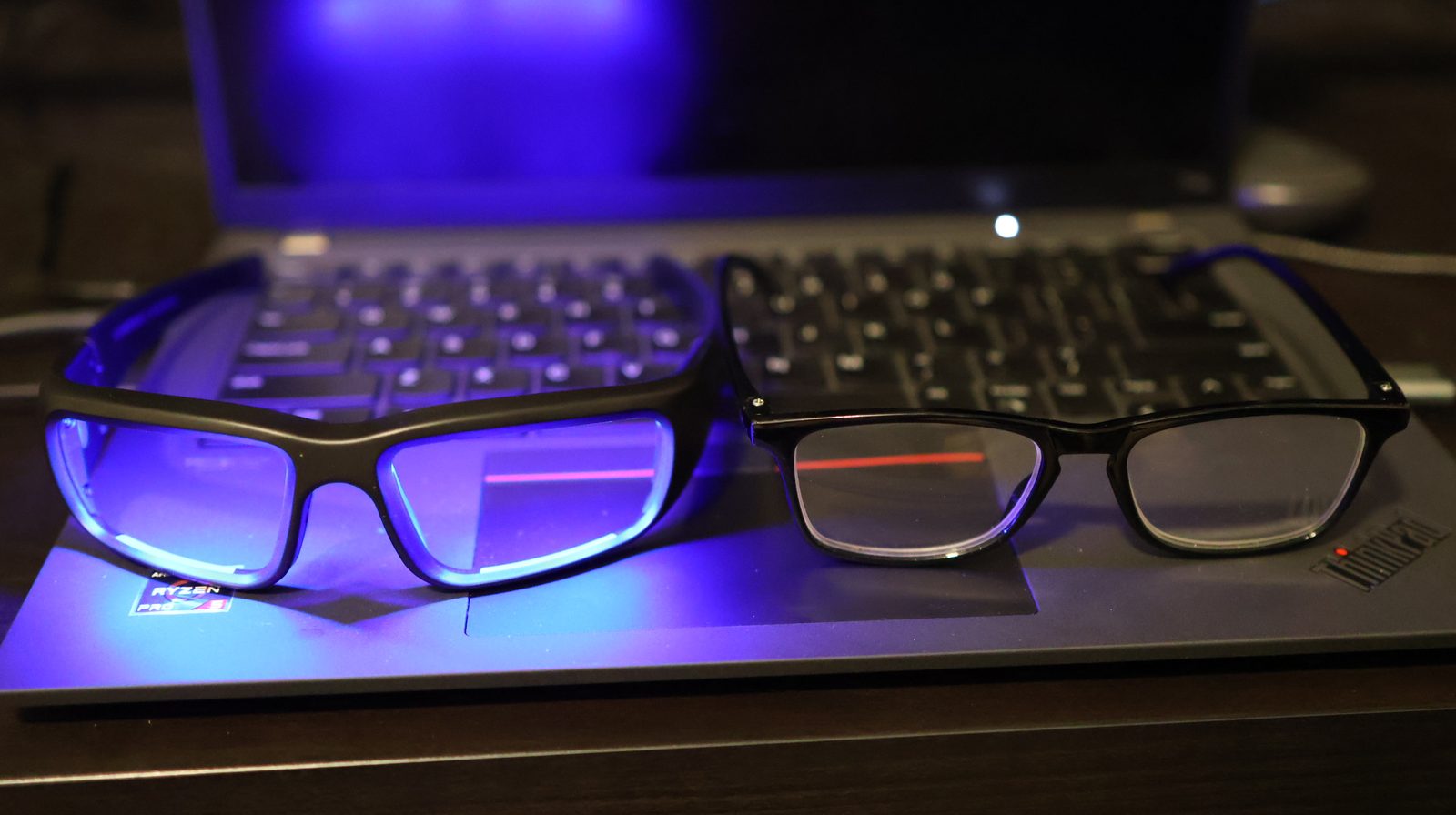 Blue light blocking gaming glasses beside clear reading glasses on a Lenovo ThinkPad laptop with AMD Ryzen Pro sticker