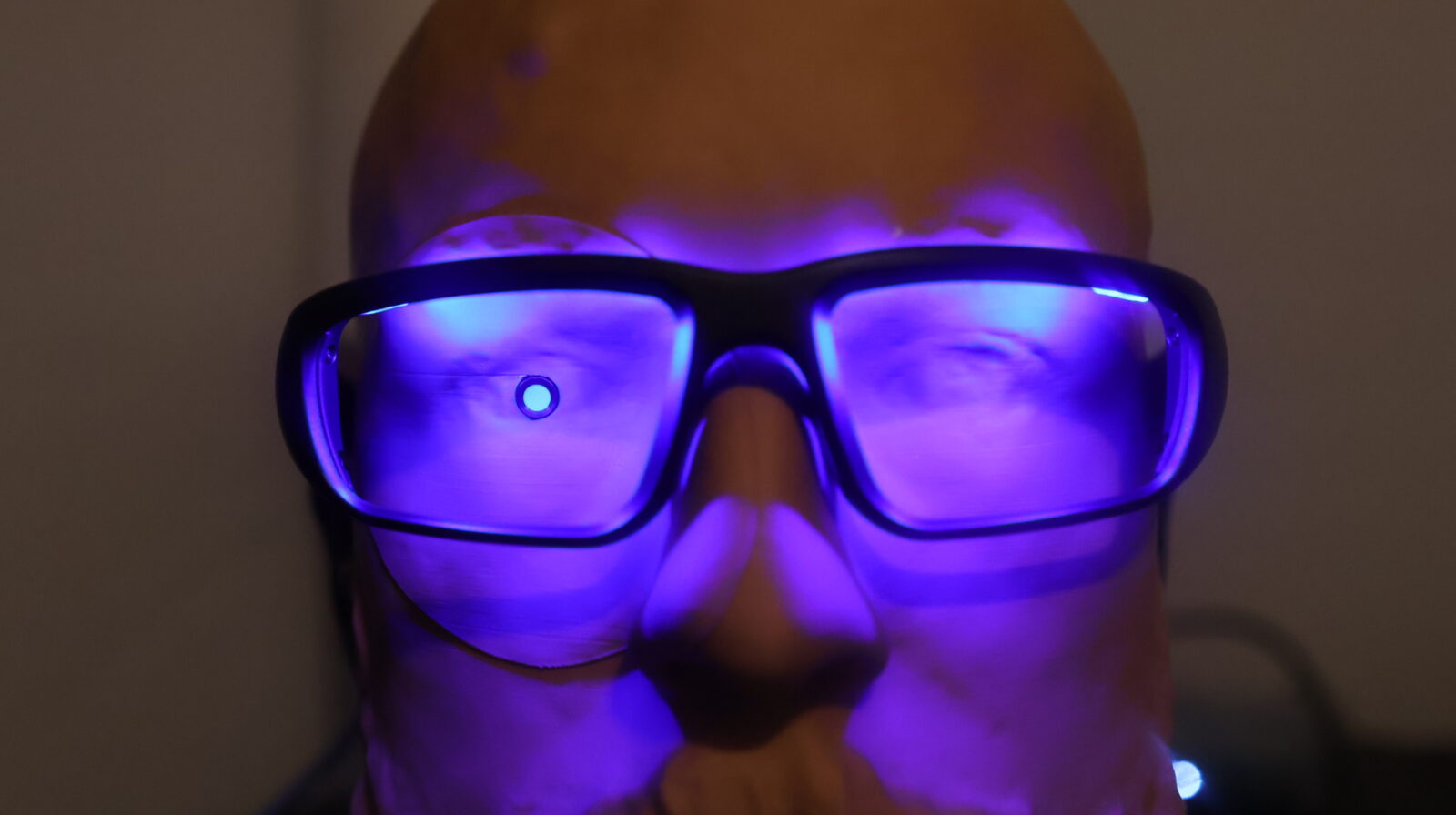 Bald man wearing glowing purple LED glasses illuminated by ultraviolet light in a dark room