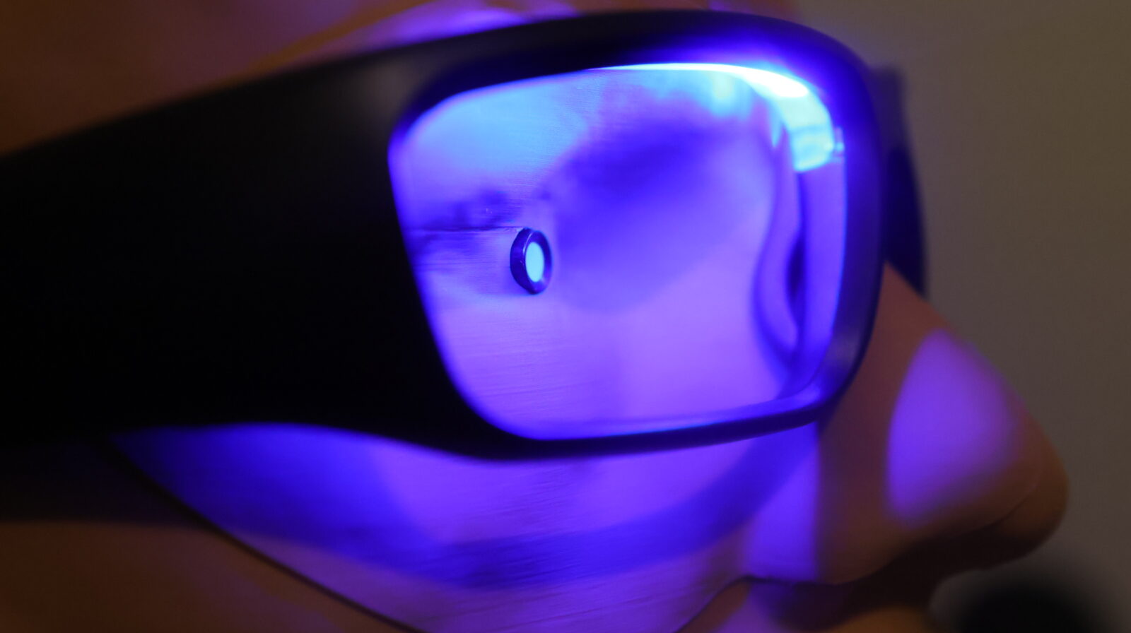 UV blacklight illuminating sunglasses lens held by hand in dark room, showing purple-blue fluorescent glow