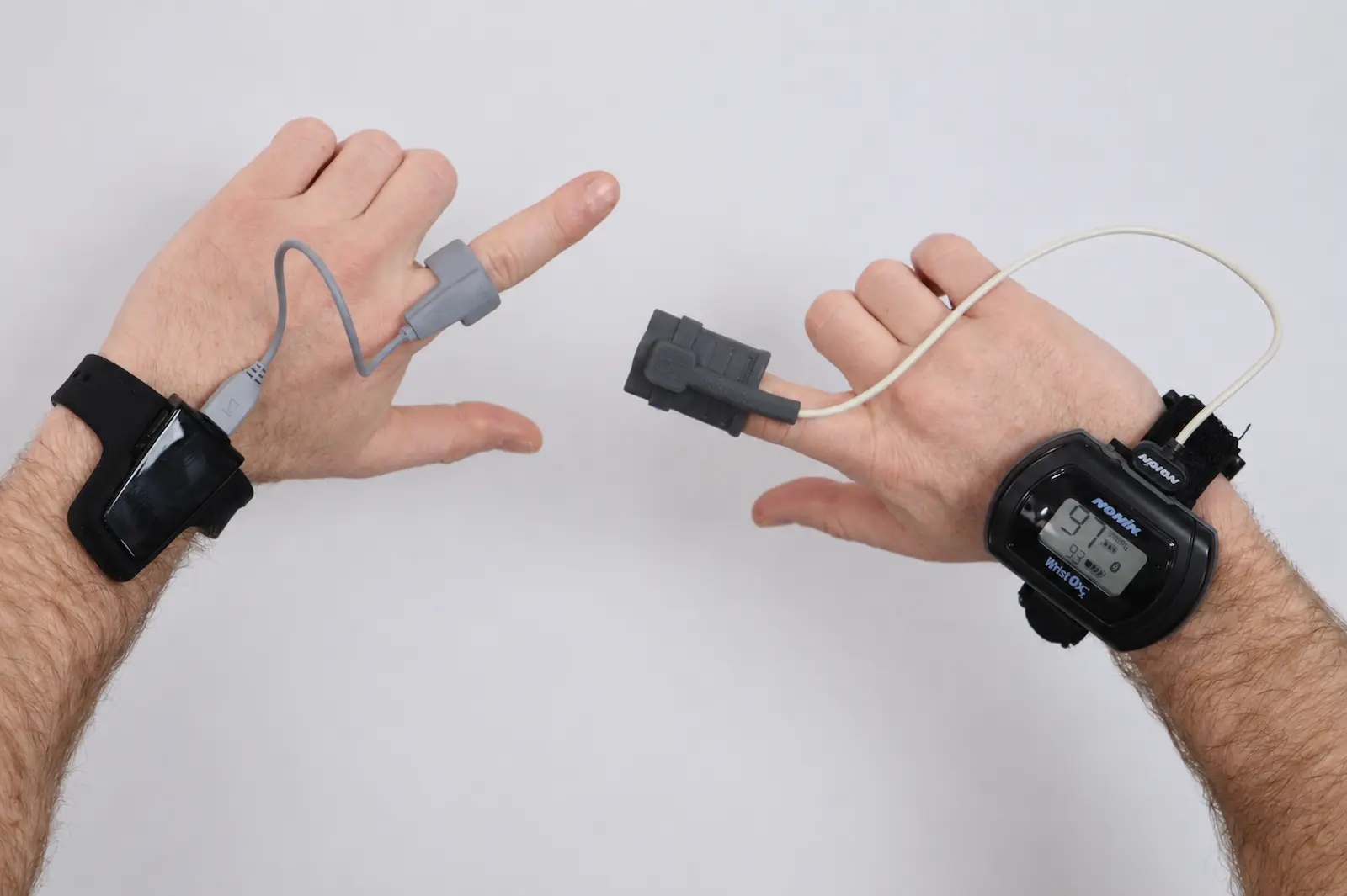 Two hands wearing pulse oximeter finger sensors and wrist monitors, including a Nonin Wrist Ox device showing SpO2 and heart