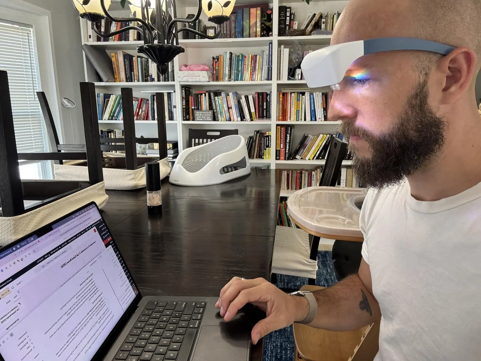 Man wearing Luminette 2 light therapy glasses while typing on a laptop at a dining table in front of bookshelves.