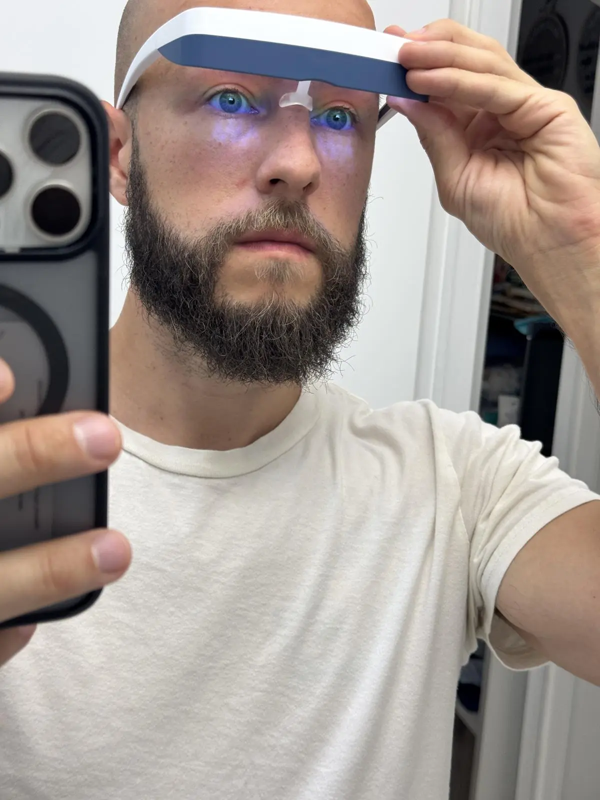 Bearded man in a white T-shirt adjusts a Luminette 3 on his nose while taking a mirror selfie, with blue light glowing under his eyes.