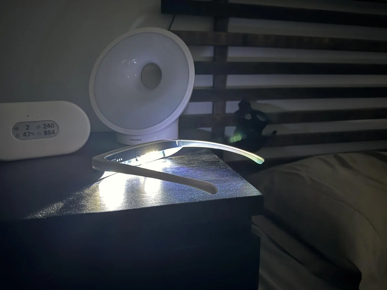 Luminette 3 glasses switched on, resting on a dark bedside table beside a white Philips light.